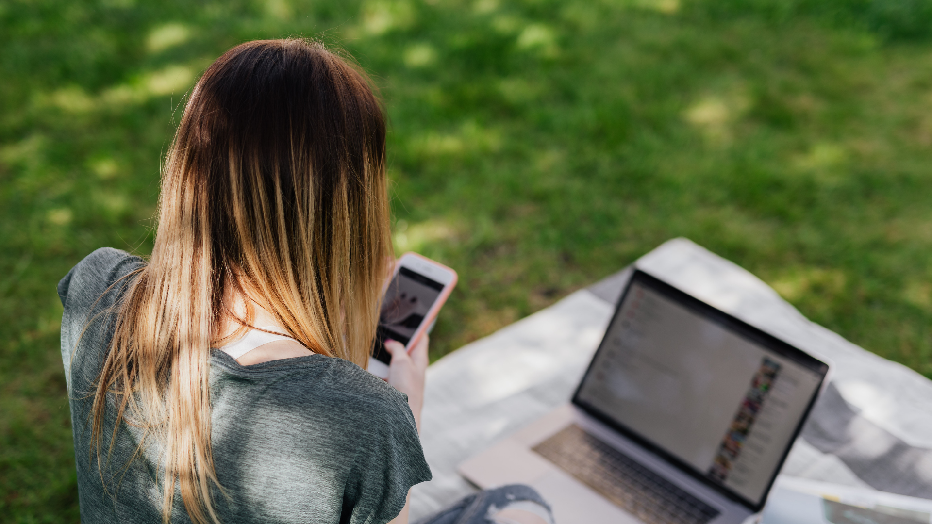 A person with long, straight hair sits on a blanket outdoors, looking at a smartphone with an open laptop beside them on the grass, working on WordPress development for Astoria Media Group.
